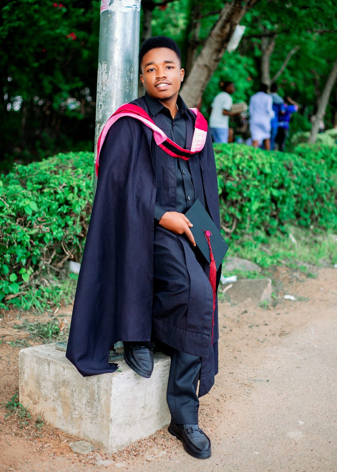 Full-length portrait of Emmanuel in graduation gown and hood, holding his cap